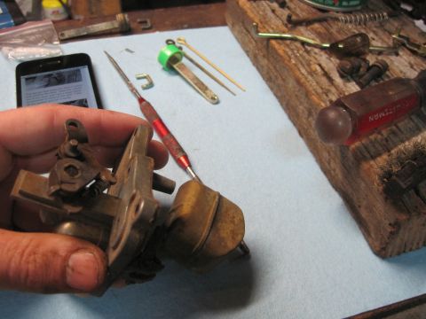 1946 Jeep Willys - A carburetor is held by a hand, with tools and a smartphone nearby, suggesting a repair or maintenance task.