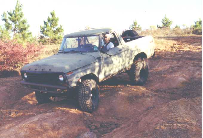 Primer-gray lifted Ford pickup with cab roof cut off crawling over red dirt off-road