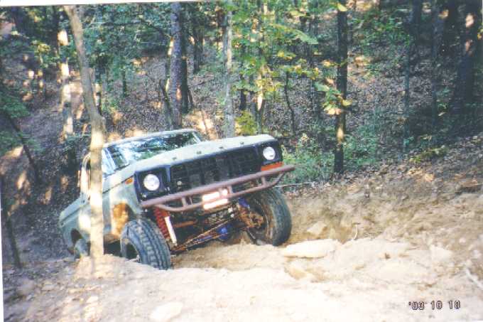Lifted green Ford Bronco with red tube bumper climbing a rocky wooded trail, front view