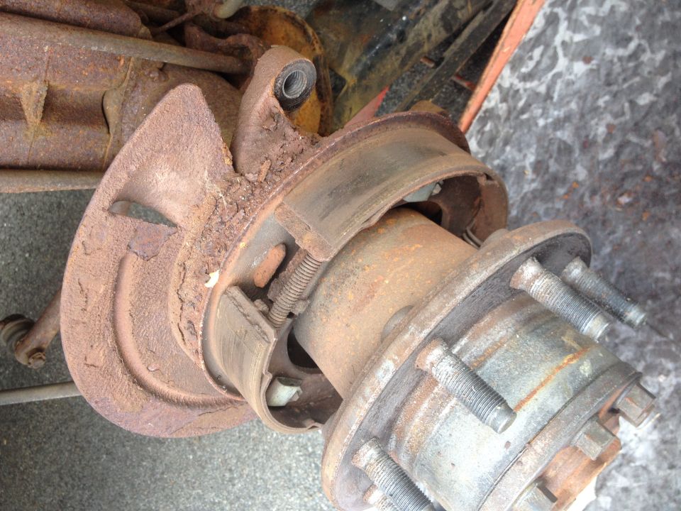 Close-up of a rusty rear axle and brake assembly with brake drum and wheel studs visible. Supermotors.net logo in lower right