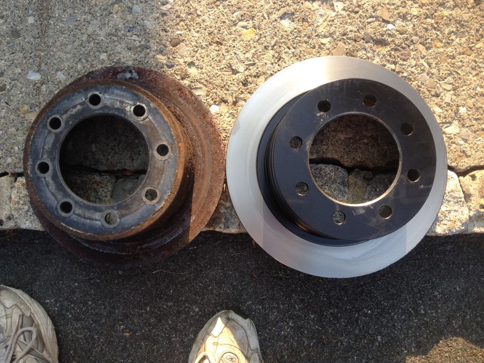 Two brake rotors, one rusty and one new, sit on asphalt