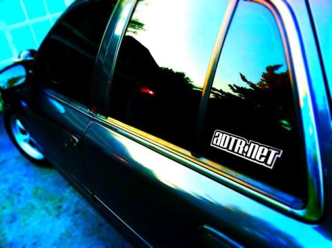 1998 Ford Crown Victoria - Rear side window of a dark sedan bearing a white 'ADTR.net' vinyl decal, with sky and trees reflected in the glass.