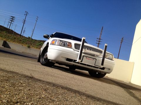 1998 Ford Crown Victoria - White Ford sedan with a front push bar and California plate, photographed from a low angle against a clear blue sky.