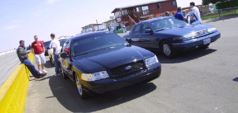 1998 Ford Crown Victoria - black Ford sedan and a dark-blue Crown Victoria parked near a building while people stand around at an event.