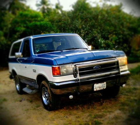 1990 Ford Bronco - This 1990 Ford Bronco is shown from a 3/4 front angle, parked on a dirt surface with trees in the background.