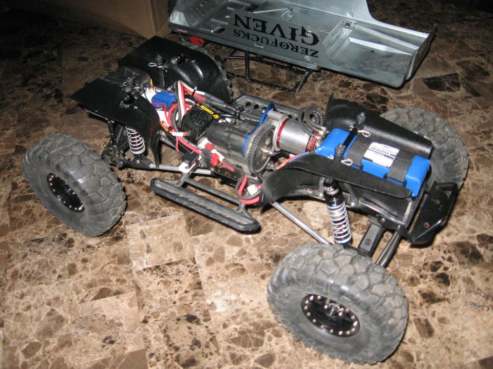 RC rock crawler chassis showing motor, battery pack, suspension, and large tires. A silver body shell with text is behind it