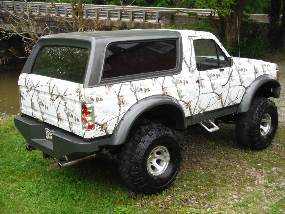 White camouflage-wrapped Ford Bronco with a grey roof and aftermarket bumper, parked on gravel and grass