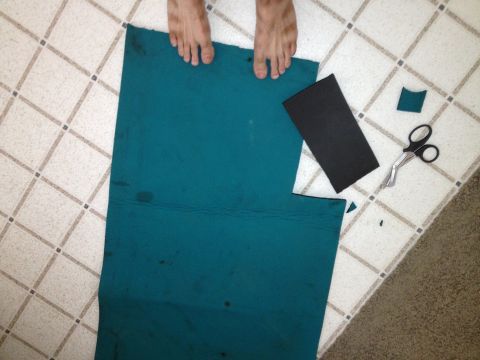 1961 International Scout 800 - A person's feet are visible as they prepare to cut teal carpet and black foam on a tiled floor with scissors.
