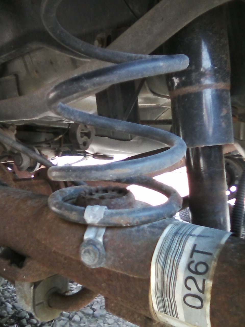 Close-up of a car's rear suspension with a coil spring and shock absorber visible. A label with text '026T' is on the axle