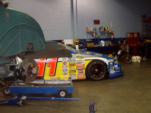 2004 Ford Crown Victoria - A race car with numerous sponsor decals is parked inside a workshop, with a brake rotor on a stand nearby.