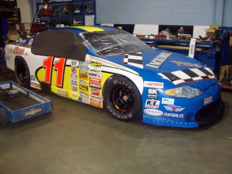 2004 Ford Crown Victoria - A race car with a blue, white, and yellow paint scheme is parked indoors next to a small cart.