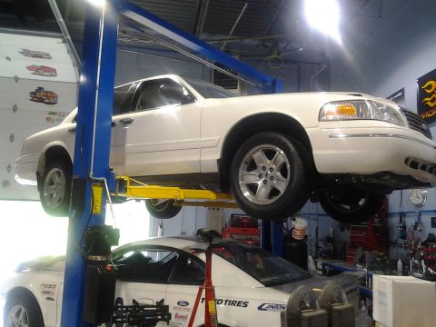 2004 Ford Crown Victoria - A white 2004 Ford Crown Victoria is elevated on a car lift in a garage, with another vehicle beneath it.