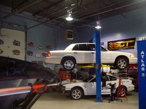 2004 Ford Crown Victoria - A white 2004 Ford Crown Victoria is elevated on a car lift in a garage, with another vehicle visible below.
