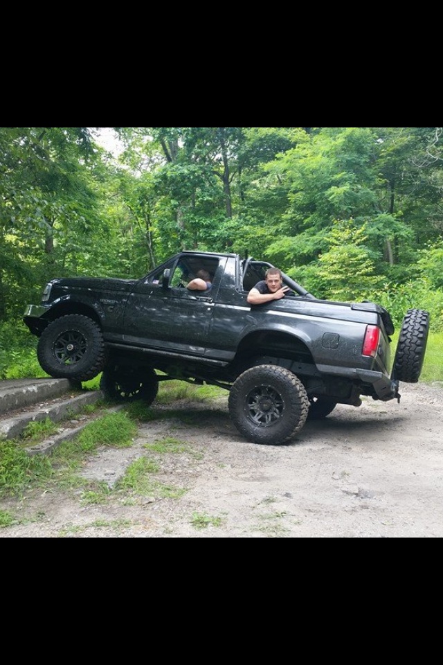Black Ford Bronco on dirt path with trees around it. Two guys are in the cab
