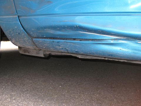 1994 Ford Thunderbird - The lower section of a blue vehicle's side panel exhibits severe damage and a detached trim piece.