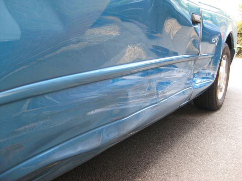 1994 Ford Thunderbird - The side of a blue car shows extensive damage, including deep dents and scraped paint.