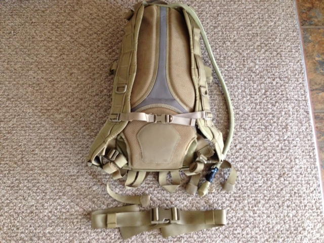 Tan tactical backpack with hydration tube and separate waist strap, lying on a carpeted floor