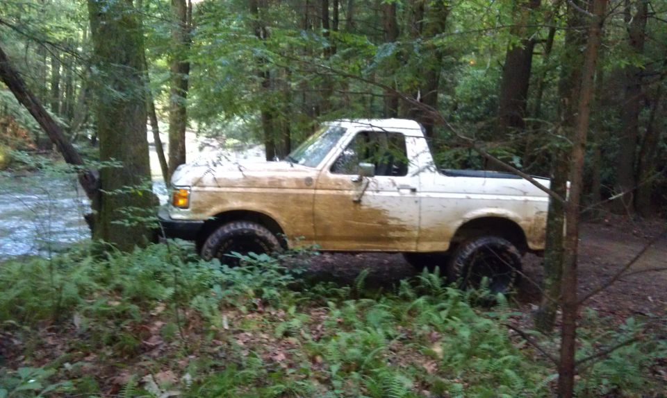 1990 Ford Bronco covered in mud, parked off-road in a forest next to a stream