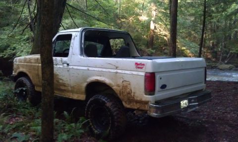 1990 Ford Bronco - A white Ford Bronco is parked off-road in a wooded area with a stream nearby.