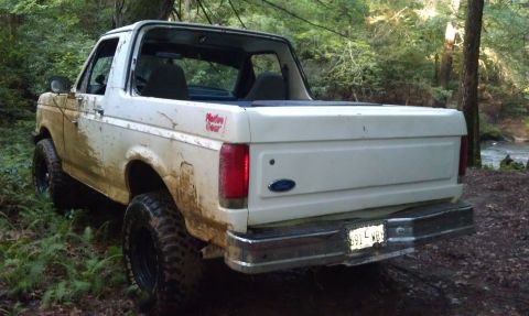 1990 Ford Bronco - Mud-covered white Ford Bronco seen off-road with a stream in the background.