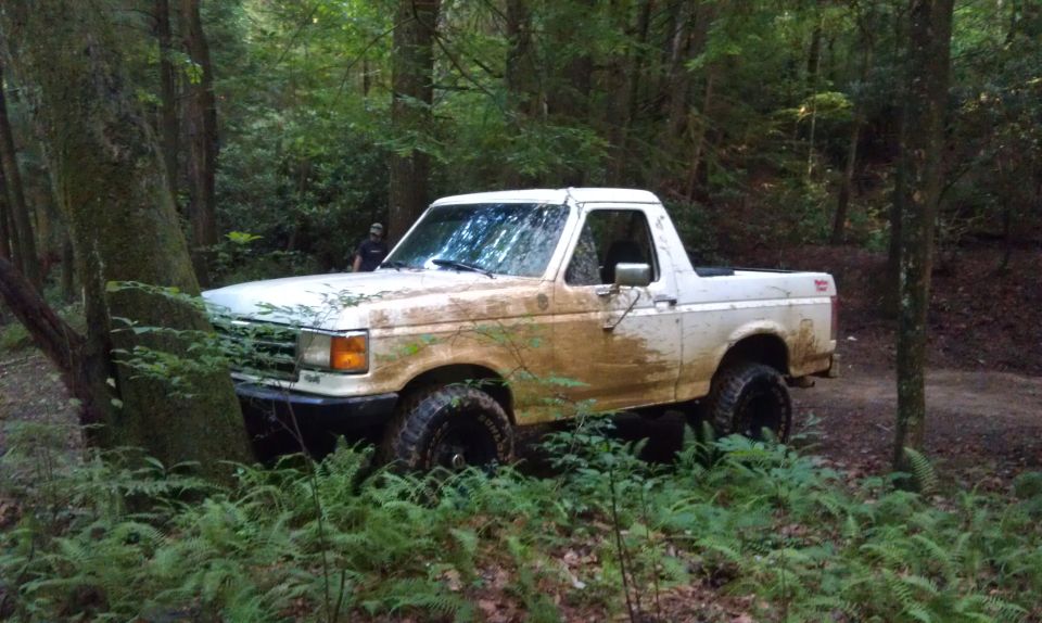 White SUV stuck in the woods, covered in mud. Someone is standing behind it