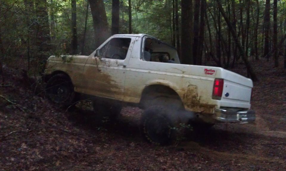 1990 Ford Bronco driving off-road on a dirt path in a wooded area, covered in mud