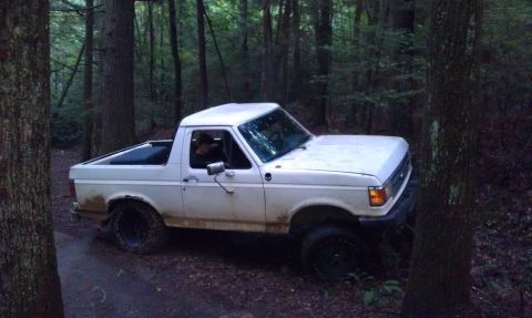 1990 Ford Bronco - A white Ford Bronco navigates a wooded trail, kicking up dust.