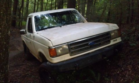 1990 Ford Bronco - A white Ford truck, likely from the late 1980s, is seen tilted off-road in a wooded area.