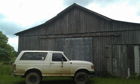 1990 Ford Bronco - A white 1987 Ford Bronco sits parked in front of a large, rustic wooden barn under a cloudy sky.