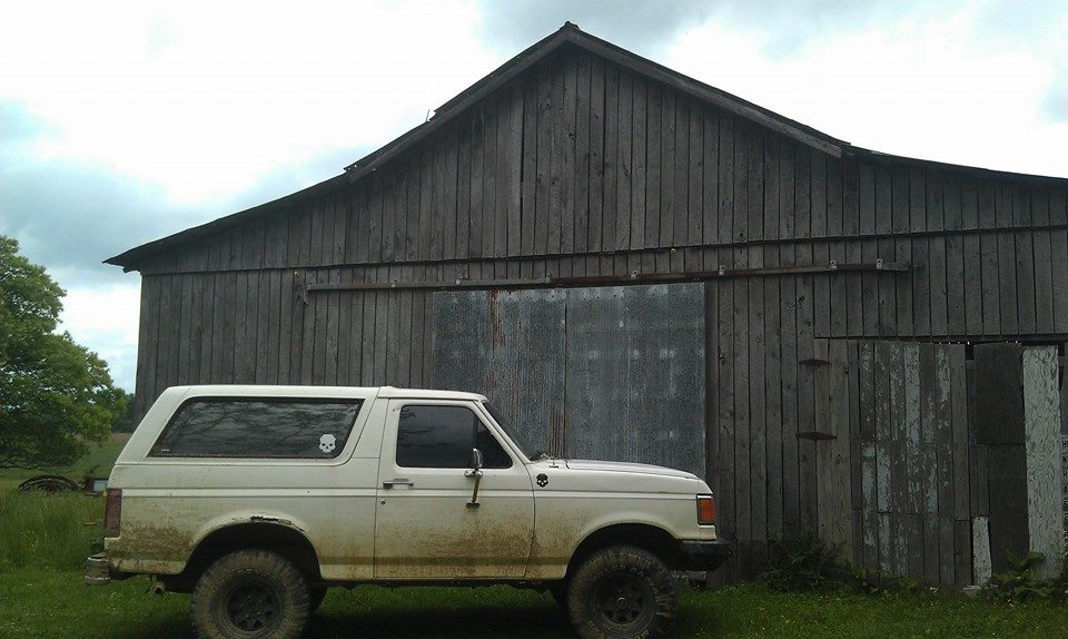 White 1987 Ford Bronco side view, muddy, parked in front of a weathered wooden barn