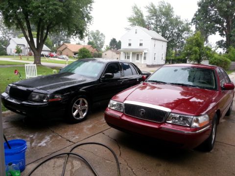 2004 Mercury Grand Marquis - Two sedans, one red and one black, are parked on a wet driveway in front of houses.