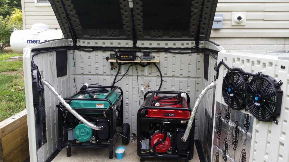Two portable generators, one green and one red, are housed inside an open plastic shed with fans mounted on the wall
