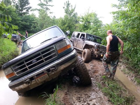 1996 Ford Bronco - Two off-road vehicles are navigating a muddy trail, with one truck deeply stuck in the mire.