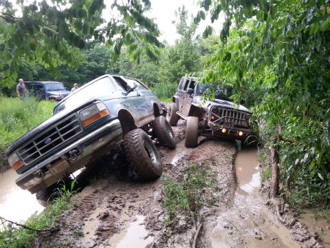 1996 Ford Bronco - Two off-road vehicles navigate a muddy trail, with one Ford Bronco and a Jeep Wrangler deeply stuck in the mire.