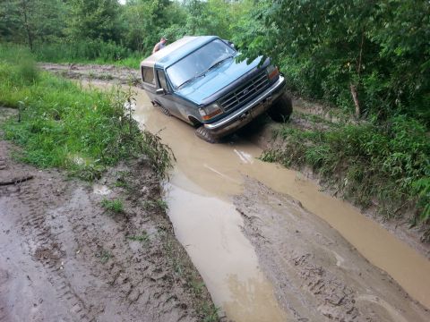 1996 Ford Bronco - A teal Ford Bronco is stuck in a muddy off-road trail with trees and foliage surrounding it.