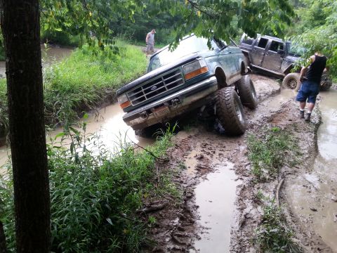 1996 Ford Bronco - Two off-road vehicles are stuck in a muddy creek bed, with one truck tilted precariously.