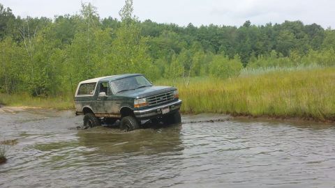 1996 Ford Bronco - A 1996 Ford Bronco navigates through a muddy, water-filled terrain surrounded by lush greenery.