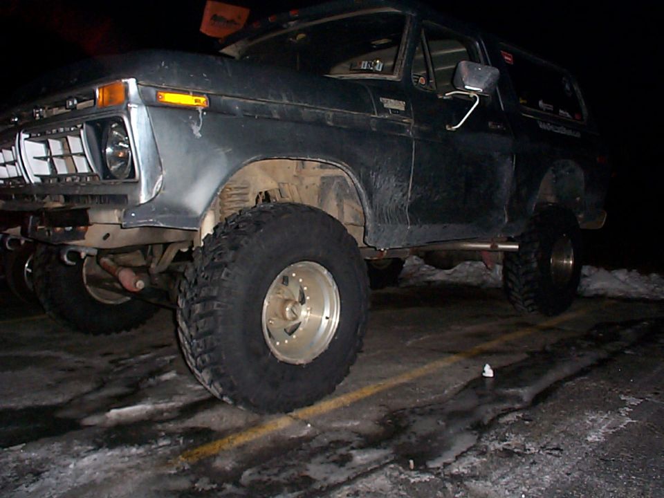 Dark colored Ford Bronco with large tires parked on a wet surface at night