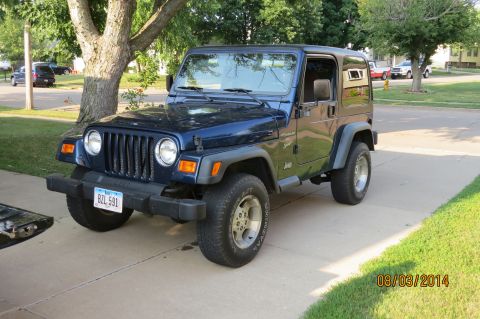 2000 Jeep Wrangler - A blue Jeep Wrangler is parked on a concrete driveway in front of a tree and residential buildings.