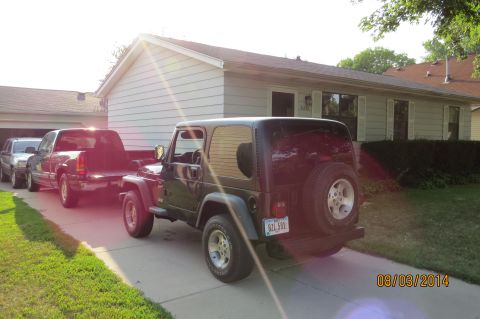 2000 Jeep Wrangler - A dark green 2000 Jeep Wrangler is parked in a driveway, with two trucks behind it.