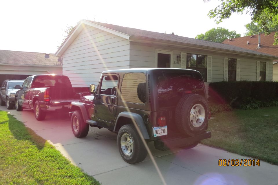 Dark green 2000 Jeep Wrangler, rear view, parked in driveway with trucks behind it