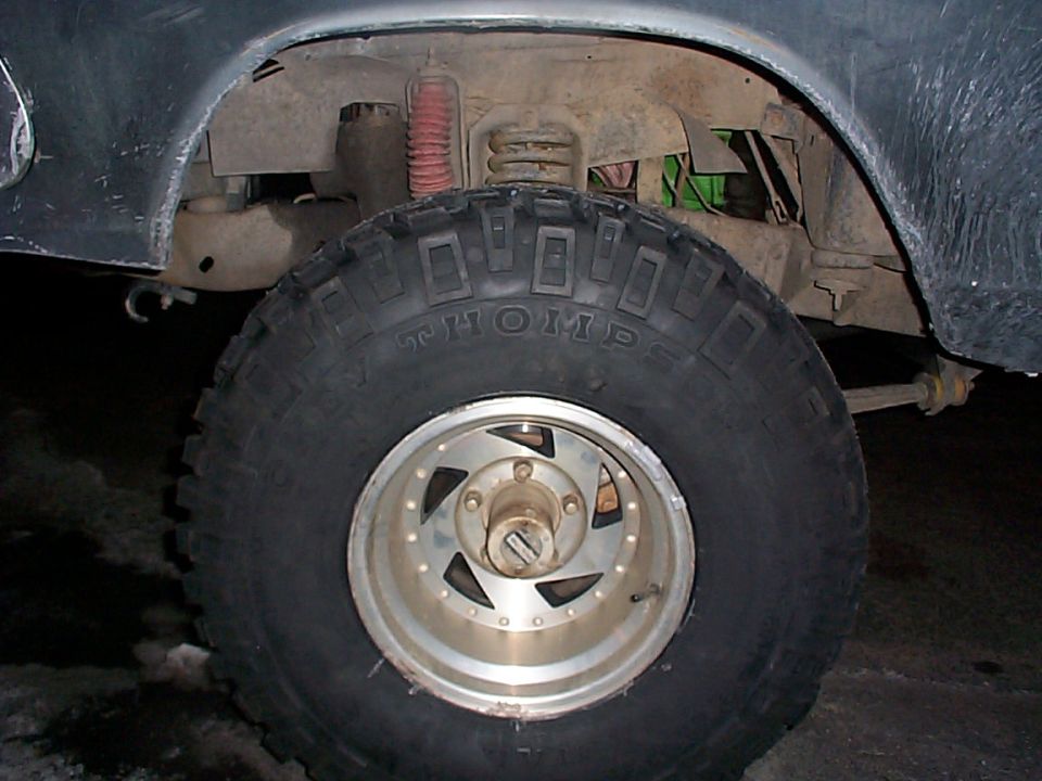 Close-up of a large off-road tire with 'THOMP' visible on the sidewall, mounted on a multi-spoke wheel. Suspension components and part of a vehicle's fender are also visible