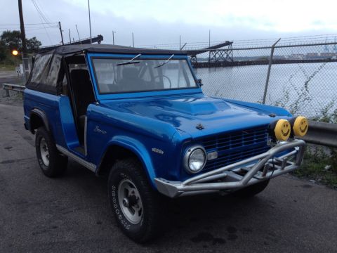 1974 Ford Bronco - A blue 1974 Ford Bronco is parked on a paved surface near a body of water under an overcast sky.