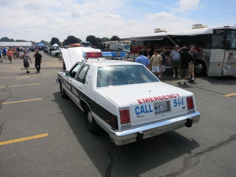 1991 Ford LTD Crown Victoria - Rear view of a white and black retired police sedan marked EMERGENCY CALL 9-1-1, parked at an outdoor show near a bus.