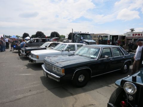 1991 Ford LTD Crown Victoria - dark blue full-size sedan leads a row of cars and emergency trucks parked on pavement at a sunny outdoor show.