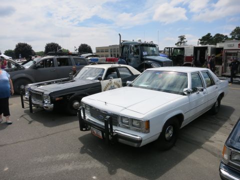 1991 Ford LTD Crown Victoria - white police sedan with a front push bar sits beside black-and-white cruisers and emergency trucks at an outdoor show.