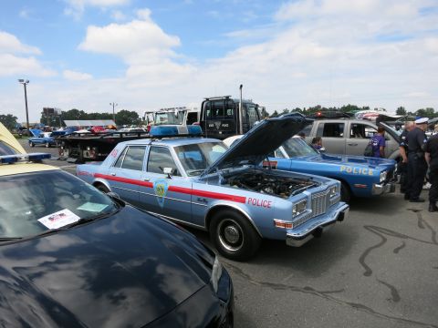 1991 Ford LTD Crown Victoria - light blue police sedan with a red stripe and open hood sits among other cruisers and a tow truck at an outdoor show.