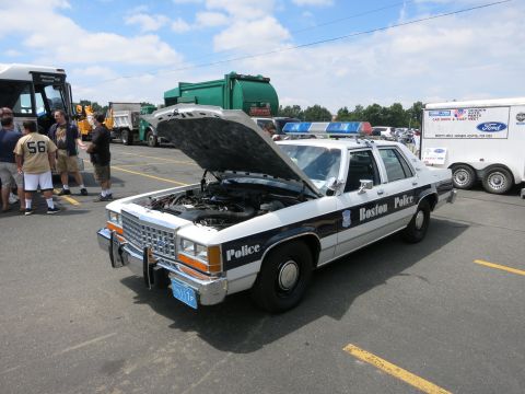 1991 Ford LTD Crown Victoria - white-and-black sedan lettered Boston Police with hood raised and front push bar sits among trucks at an outdoor event.