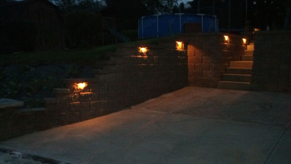 Backyard patio with stone steps and lights at night. Blue pool in background