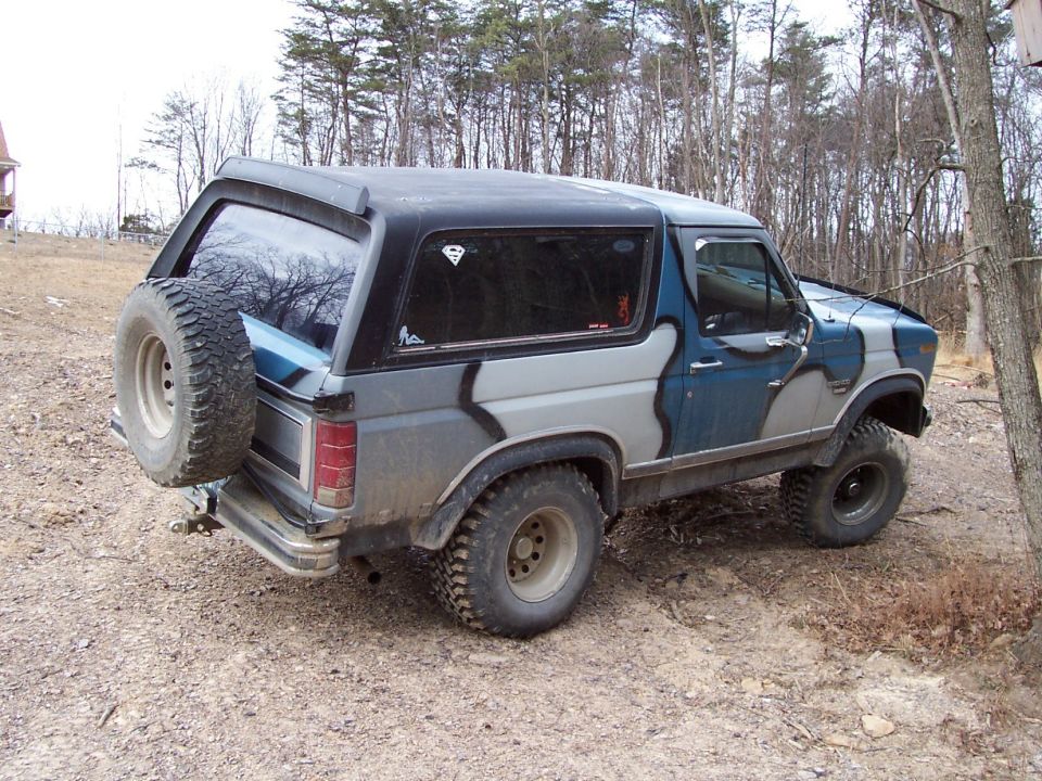 Ford Bronco with camouflage paint, spare tire on rear door, parked on dirt road
